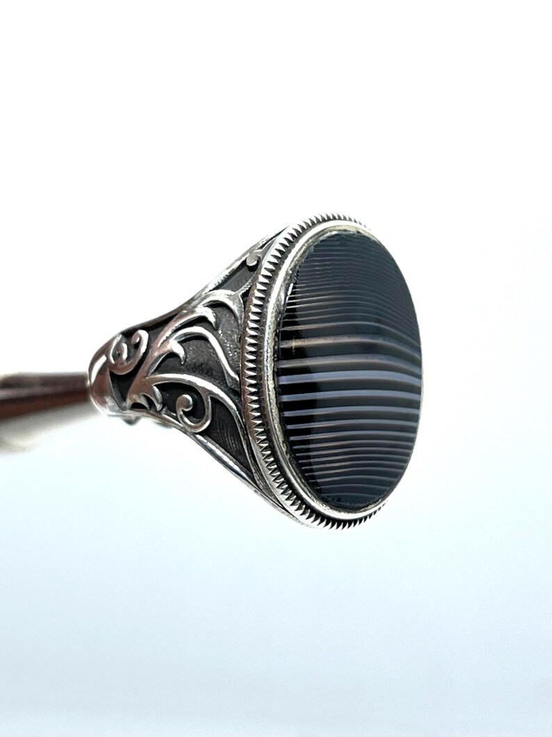 May include: A silver ring with an oval, black and gray striped stone. The ring has ornate, swirling designs on the band and around the stone setting. The stone is approximately 1 inch long.
