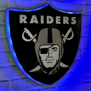 May include: A black and silver shield-shaped sign with the word "RAIDERS" in white letters and a stylized image of a football player wearing a helmet and an eye patch. The sign is illuminated with blue LED lights.
