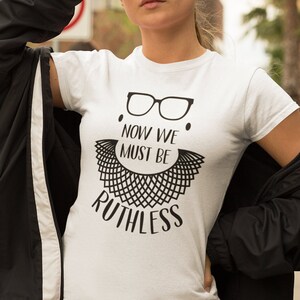 Può includere: T-shirt bianca con grafica nera di occhiali, colletto in pizzo e la scritta "NOW WE MUST BE RUTHLESS". La maglietta è a maniche corte.