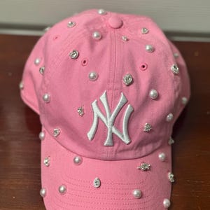 May include: Pink baseball cap with white embroidered "NY" logo and embellished with faux pearls and rhinestones. The cap has a curved brim and a button on top. A stylish accessory.