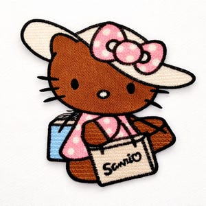Peut inclure: Écusson brodé représentant un chat de dessin animé marron portant un chapeau blanc avec un nœud rose à pois. Le chat porte une robe rose à pois et porte des sacs de courses. Le sac de courses porte le mot "Sanrio".