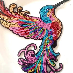 Peut inclure: Applique de colibri orné de sequins colorés. L'oiseau présente un contour noir et est orné de sequins bleus, roses, rouges, jaunes et verts. L'applique est conçue avec des ailes détaillées et une longue queue, idéale pour embellir des vêtements ou des accessoires.
