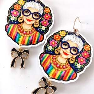 Peut inclure: Une paire de boucles d'oreilles fantaisistes représentant une femme avec des lunettes, un haut rayé arc-en-ciel et une couronne de fleurs. Chaque boucle d'oreille a un charme en forme de nœud noir. Les boucles d'oreilles ont des crochets dorés.