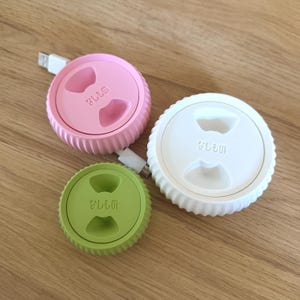 May include: Three cable organizers in pink, white, and green. Each organizer has a raised rim and a small indentation in the center. The organizers are labeled "glem".