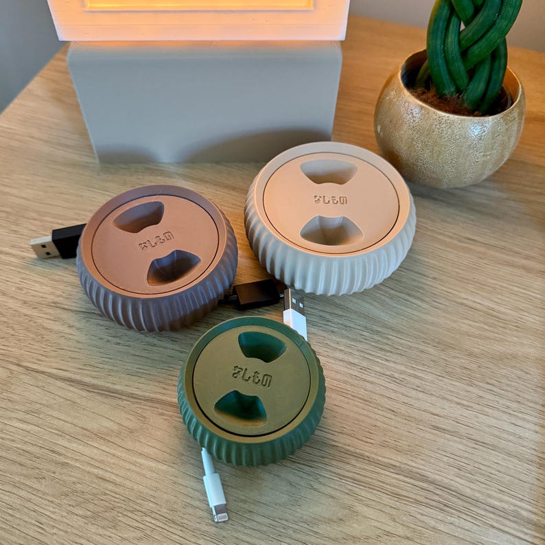 May include: Three cable organizers in different colors: brown, beige, and green. Each organizer has a circular shape with a groove in the center and the word "SL&M" printed on the top. The organizers are on a wooden surface.