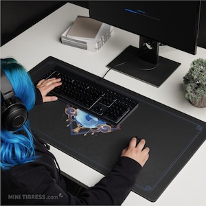 May include: Black rectangular gaming mouse pad with a blue and gold design, a black keyboard, and a black mouse. The mouse pad has a blue border and a decorative design. The image also shows a computer monitor and a small plant.