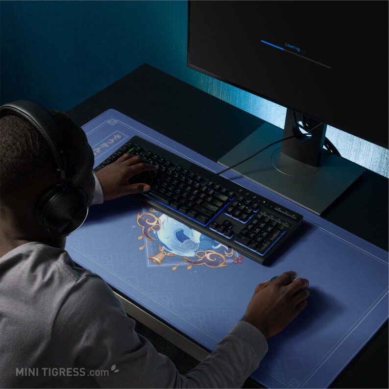 May include: A person using a black keyboard and mouse on a blue gaming mousepad. The mousepad has a decorative design with gold accents. A computer monitor displays a loading screen. The website address "MINI TIGRESS.com" is visible.