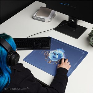 May include: A blue rectangular mousepad with a stylized blue and white graphic design. The mousepad is on a white desk next to a black keyboard and a computer monitor. The mousepad has a decorative border.
