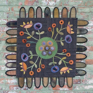May include: A colorful wool applique wall hanging with a floral design. The design features a central circle with a green background and blue and orange circles. The circle is surrounded by stylized flowers with orange, blue, and green leaves. The wall hanging is framed by a border of brown and tan fabric.
