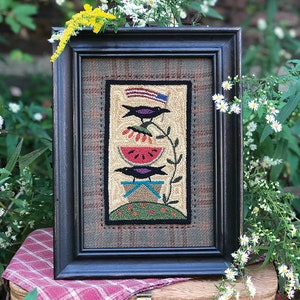 May include: A framed needlepoint artwork depicting a black bird perched on a watermelon slice with a red, white, and blue flag in the background. The artwork is framed in a black wood frame and has a plaid background.