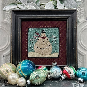 May include: A framed needlepoint design of a snowman wearing a black top hat and a red scarf, holding a string of white beads. The snowman is standing on a blue background with white snow.