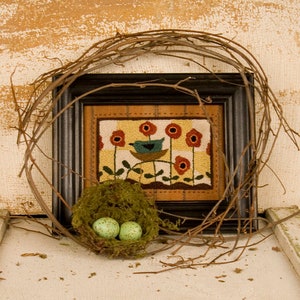 May include: A framed needlepoint artwork depicting a blue bird perched on a nest with red flowers and green leaves on a yellow background. A small bird's nest with two green eggs sits in front of the artwork.