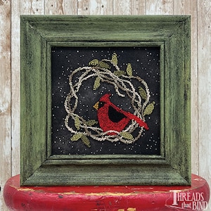 May include: A framed needlepoint artwork featuring a red cardinal perched on a green wreath against a black background with white dots. The frame is a dark green wood.