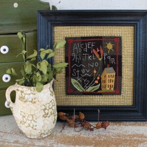 May include: A framed needlepoint piece with a black background and a tan burlap border. The needlepoint design features a small yellow house with a red roof, a red flower, and the alphabet in a black font. The text reads "ABCDEFGHIJKLMNOPQRSTUVWXYZ".