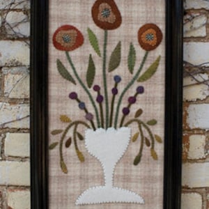 May include: A framed needlepoint artwork featuring a bouquet of orange and red flowers in a white vase. The flowers are surrounded by green leaves and purple berries. The background is a beige fabric with a subtle plaid pattern.