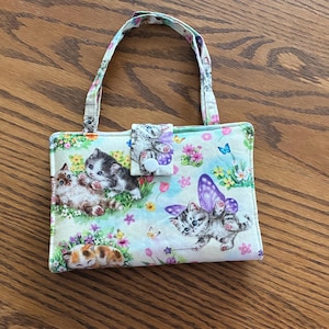 May include: A small, rectangular handbag with a handle, featuring a colorful print of kittens, butterflies, and flowers. The bag has a button closure and a light-colored background. The handle and trim have a matching floral pattern.