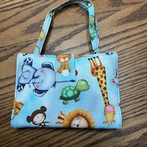 May include: A small, light blue handbag with a repeating pattern of cartoon animals, including elephants, giraffes, and monkeys. The bag has a matching handle and a button closure. The background is a light blue color.