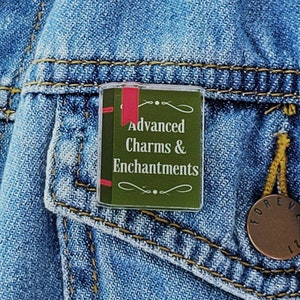May include: A green and red enamel pin shaped like a book with the title "Advanced Charms & Enchantments" on a denim jacket.