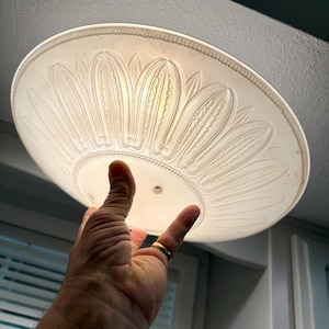 May include: A circular, translucent white glass light fixture shade featuring a molded, raised petal or leaf pattern radiating from the center. The edge of the shade has a decorative beaded border, and a small circular hole is visible in the center.