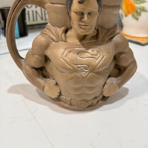 May include: A tan ceramic mug shaped like the torso of Superman, featuring a sculpted chest with the iconic S-shield emblem, a cape detail on the shoulders, a belt at the waist, and a dark blue rim at the top with a curved handle on the left side.