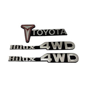 Może przedstawiać: Trzy emblematy samochodowe. Górny emblemat głosi "TOYOTA" srebrnymi literami na czarnym tle, z czerwonym i srebrnym logo. Poniżej dwa emblematy "Hilux 4WD" w kolorze srebrnym na czarnym.