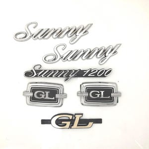 May include: A set of six silver and black car emblems. The emblems include the words "Sunny" and "Sunny 1200" and the letters "GL".