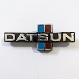 May include: A black and silver Datsun emblem with a red and blue stripe design.