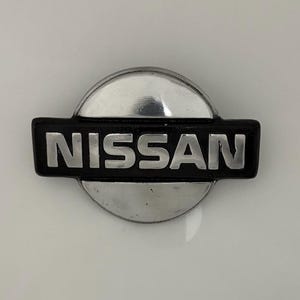 Puede incluir: Un emblema de Nissan plateado y negro con la palabra "NISSAN" en letras plateadas.
