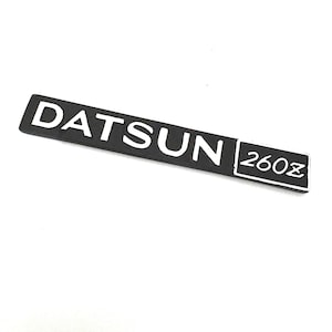 Könnte beinhalten: Schwarzweißes Datsun 260Z Emblem. Das Emblem ist rechteckig mit abgerundeten Ecken und trägt den Schriftzug "Datsun" in weißen Buchstaben auf schwarzem Hintergrund. Die Zahl "260Z" befindet sich in weißen Buchstaben auf schwarzem Hintergrund in einem kleineren Rechteck rechts neben dem Wort "Datsun".