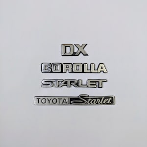 May include: Silver and black car emblems with the text "DX", "COROLLA", "STARLET", and "TOYOTA Starlet".