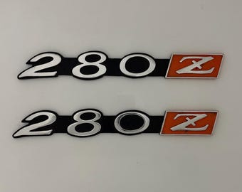 May include: Two 280Z car emblems. Each emblem has the numbers "280" in silver with a black background, and an orange rectangle with a silver "Z" on the right side. The emblems are on a white background.