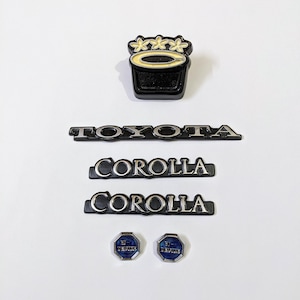 Könnte beinhalten: Ein Satz schwarzer und silberner Toyota Corolla Autoembleme. Das Set enthält ein schwarzes und silbernes Toyota Emblem, zwei schwarze und silberne Corolla Embleme und zwei blaue und silberne "EFI" Embleme.