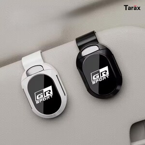 May include: Two oval-shaped car visor sunglass clips, one in white and one in black, each featuring a glossy centre panel with the white text GR SPORT printed on it. The clips have a spring-loaded mechanism and are attached to a textured surface.
