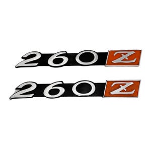 May include: Two silver 260Z emblems with black backgrounds and orange Zs. The emblems are for a car.