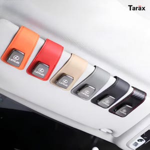 Puede incluir: Seis soportes para gafas de sol para visera de coche en naranja, rojo, beige, gris, negro y patrón de fibra de carbono, cada uno con un clip metálico plateado con el logo de Lexus. Estos accesorios están sujetos a una visera clara con el texto Tarax.