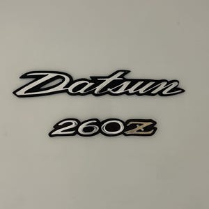 May include: A silver Datsun emblem with black trim and a silver 260Z emblem with a gold Z.