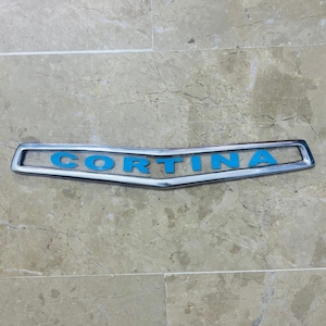 Może przedstawiać: Srebrny emblemat samochodowy z napisem "Cortina" w niebieskich literach.