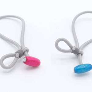 Op de afbeelding: Twee grijze elastische lussen met een knoop aan het einde. Eén lus heeft een roze kraal en de andere een blauwe kraal.