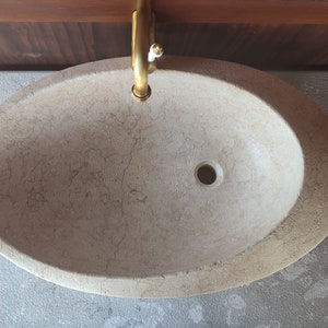 Op de afbeelding: Een beige ovale badkamer wastafel met een afvoergat in het midden. De wastafel is gemaakt van natuursteen met een marmerpatroon.