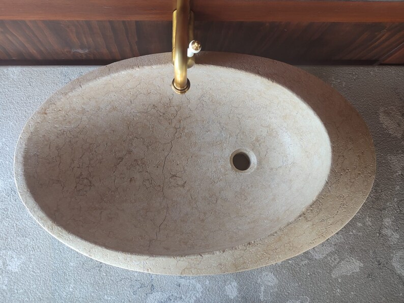 Op de afbeelding: Een beige ovale badkamer wastafel met een afvoergat in het midden. De wastafel is gemaakt van natuursteen met een marmerpatroon.
