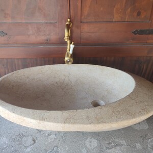 Op de afbeelding: Een grote, ovale, lichtbeige stenen wastafel met een afvoergat. De wastafel staat op een lichtgrijs aanrechtblad. Een gouden kraan is aan de muur achter de wastafel gemonteerd.