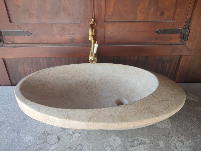 Op de afbeelding: Een grote, ovale, lichtbeige stenen wastafel met een afvoergat. De wastafel staat op een lichtgrijs aanrechtblad. Een gouden kraan is aan de muur achter de wastafel gemonteerd.