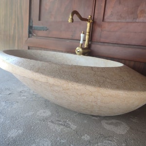 Op de afbeelding: Een grote, ovale, lichtbeige stenen wastafel met een gouden kraan. De wastafel staat op een grijs aanrecht.