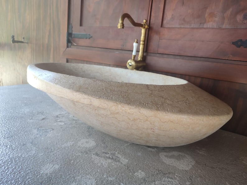 Op de afbeelding: Een grote, ovale, lichtbeige stenen wastafel met een gouden kraan. De wastafel staat op een grijs aanrecht.