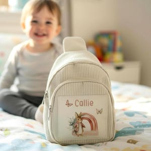 Puede incluir: Una mochila beige de pana con un bolsillo frontal con cremallera. La mochila presenta un parche rectangular con el nombre "Callie", una ilustraci&oacute;n de acuarela de un conejo, un arco&iacute;ris y mariposas. La mochila est&aacute; dise&ntilde;ada para un ni&ntilde;o.
