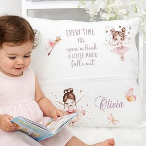 Op de afbeelding: Een wit kussen met een zak, versierd met een ballerina-ontwerp en de tekst "Every time you open a book a little magic falls out." Het kussen heeft ook de naam "Olivia" erop gedrukt. Een jong kind houdt een boek vast.