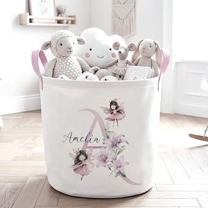 May include: A white fabric toy storage basket with pink handles, featuring the name "Amelia" and a floral and fairy design. The basket is filled with stuffed animals, including a lamb, cloud, mouse, and bunny.