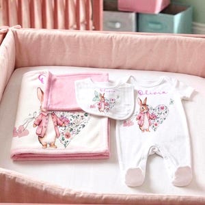 May include: A white baby blanket, a small bib, and a white footed onesie, all featuring a printed illustration of a rabbit wearing a pink jacket next to a floral heart. The name Olivia is printed in pink script on the onesie and the bib.