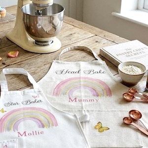 May include: White aprons with rainbow designs and the text "Star Baker" and "Head Bake Mummy". A stand mixer, measuring spoons, and a cookbook are on a wooden surface. Vanilla extract and flour are also visible, alongside baking equipment.