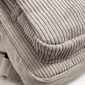 Puede incluir: Primer plano de una mochila de pana beige. La tela tiene una textura acanalada, con l&iacute;neas verticales visibles. El dise&ntilde;o de la bolsa incluye una solapa y una esquina redondeada, lo que sugiere un estilo informal. El color es un marr&oacute;n claro neutro.